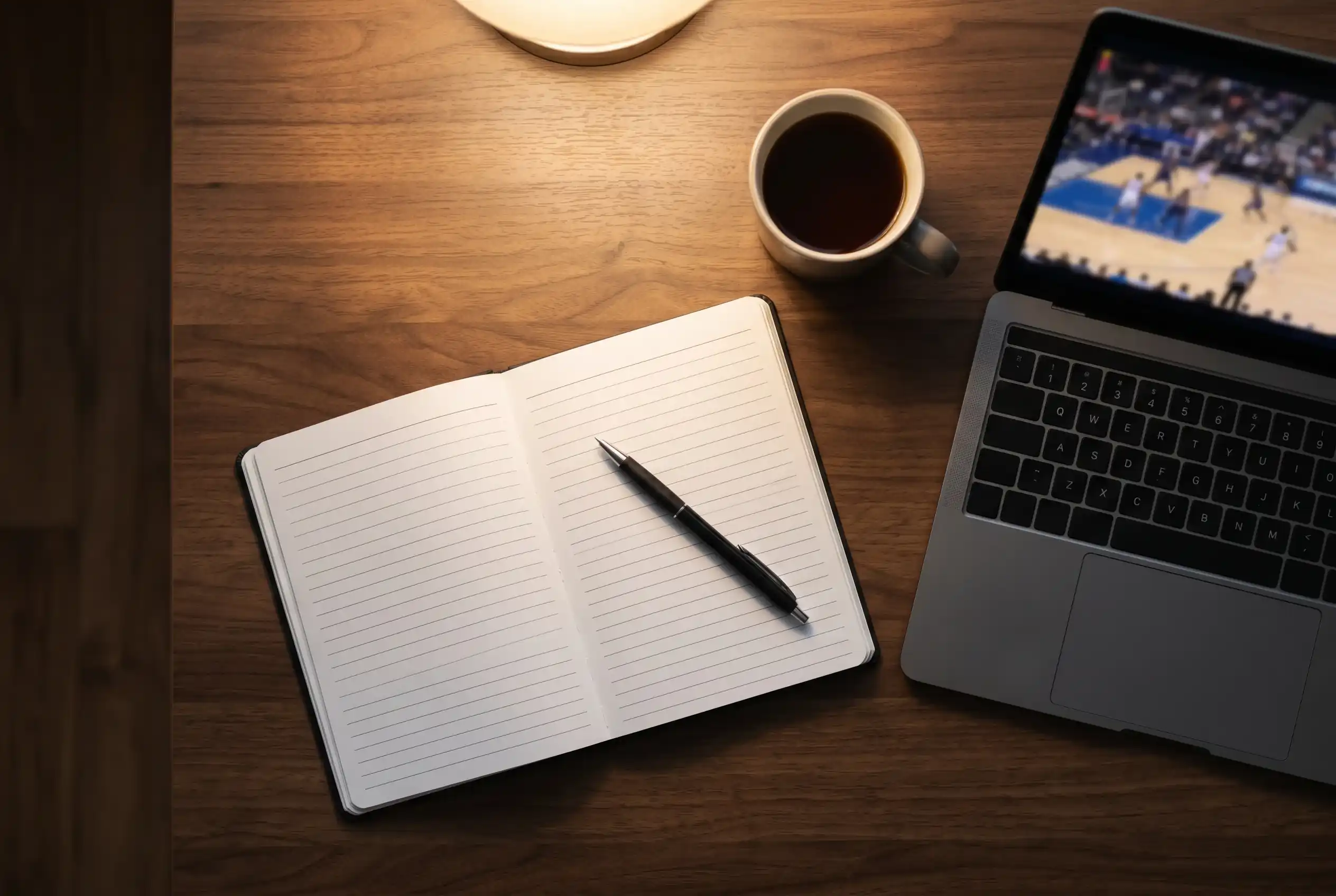 Notebook and pen on a desk beside a laptop showing a basketball match representing disciplined bankroll management